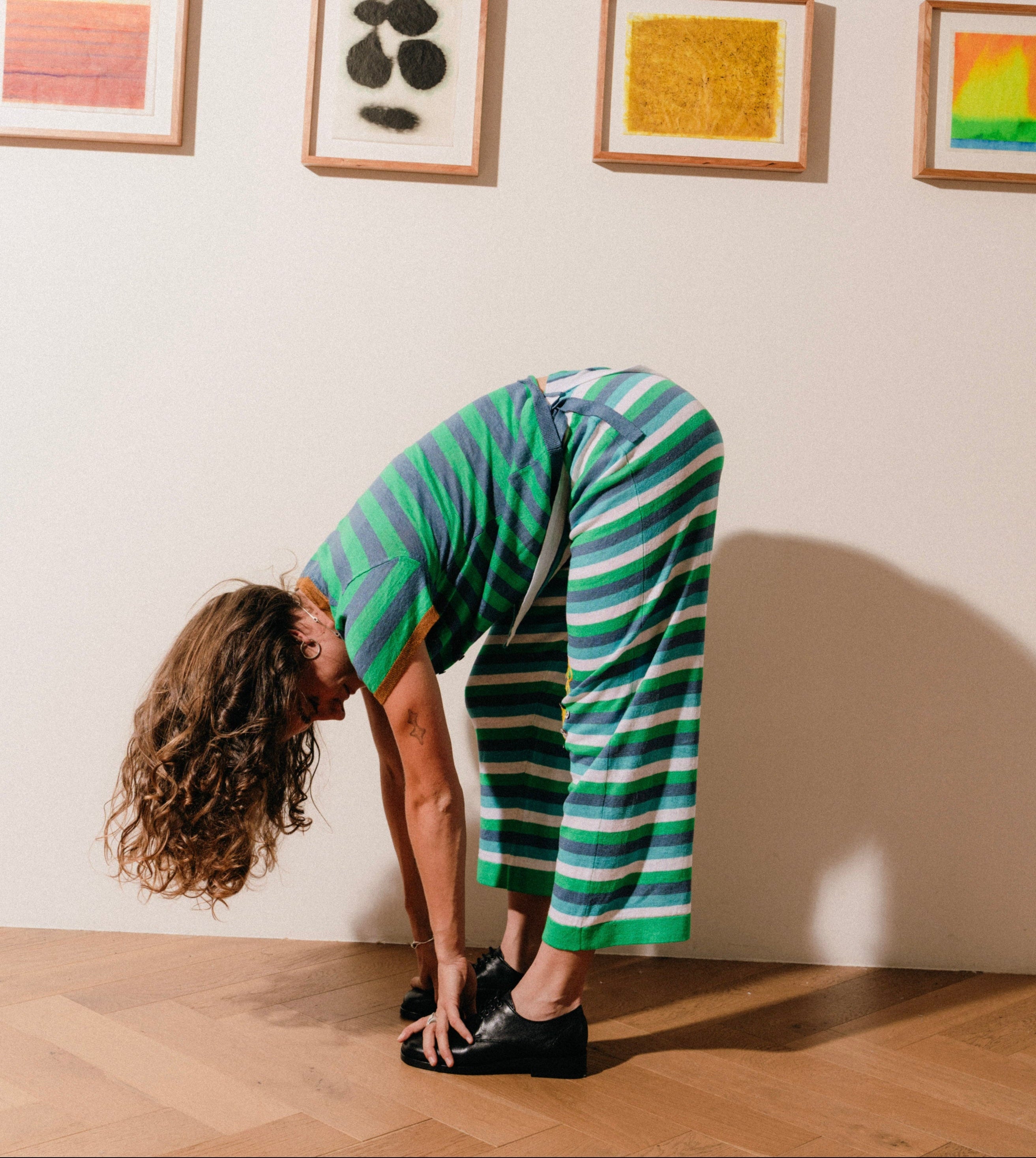 Person in a green and blue striped outfit bending over in front of framed artworks on a wall.