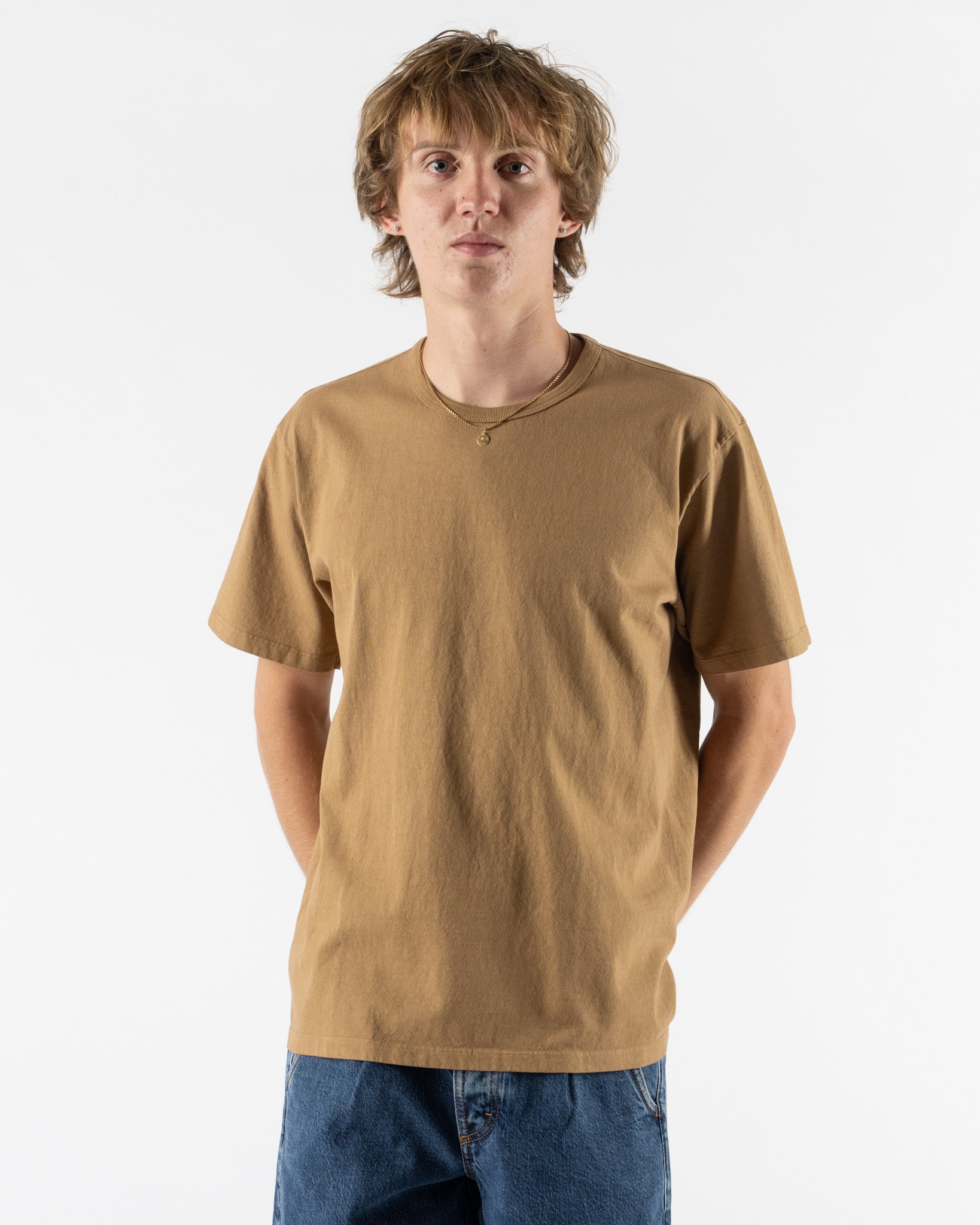 A young man with light brown hair wearing a tan short-sleeve crewneck t-shirt and blue jeans, standing against a plain white studio background.